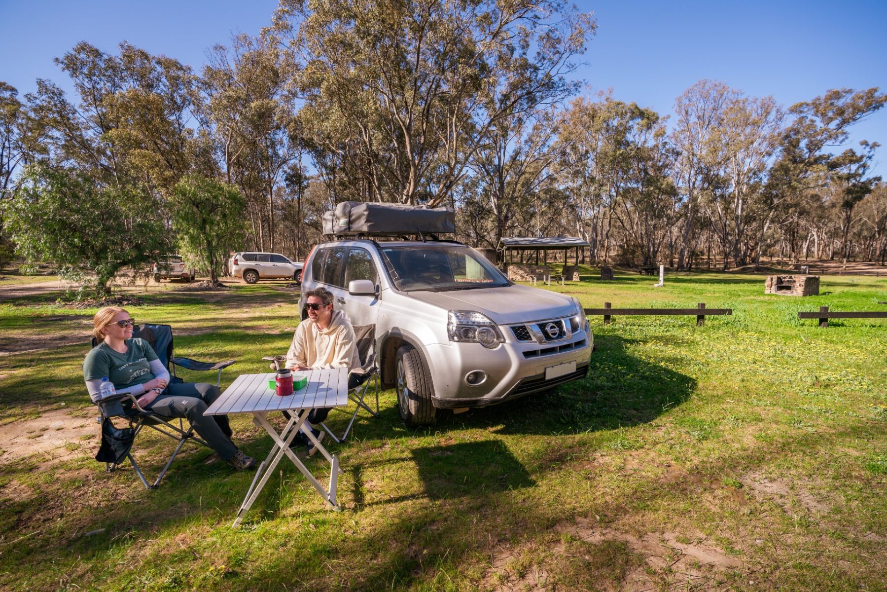 A woman and a man sitting at a small table next to a 4WD vehicle on the grasss