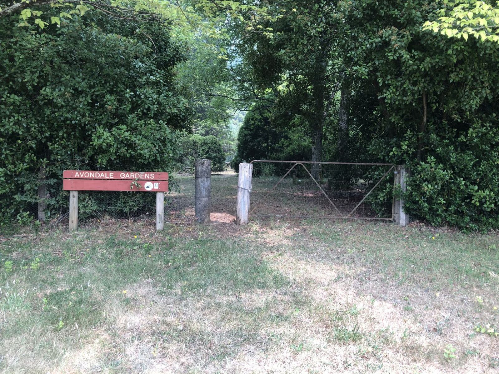A wooden sign reading Avondale Gardens next to a metal gate across a dirt pathway aligned with green, leafy trees. 