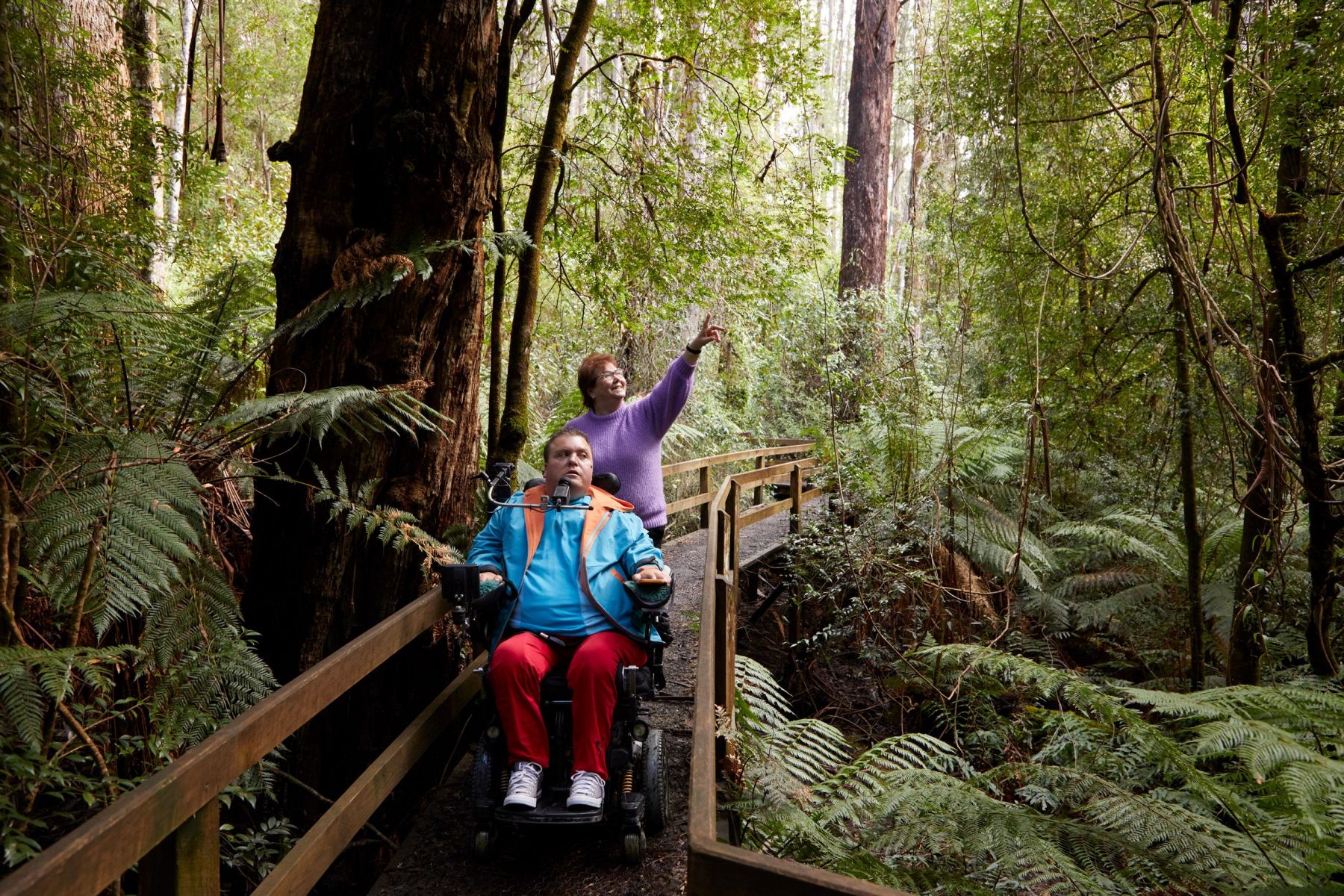 A woman pushing a man in a wheelchair along a boardwalk in the forest. 