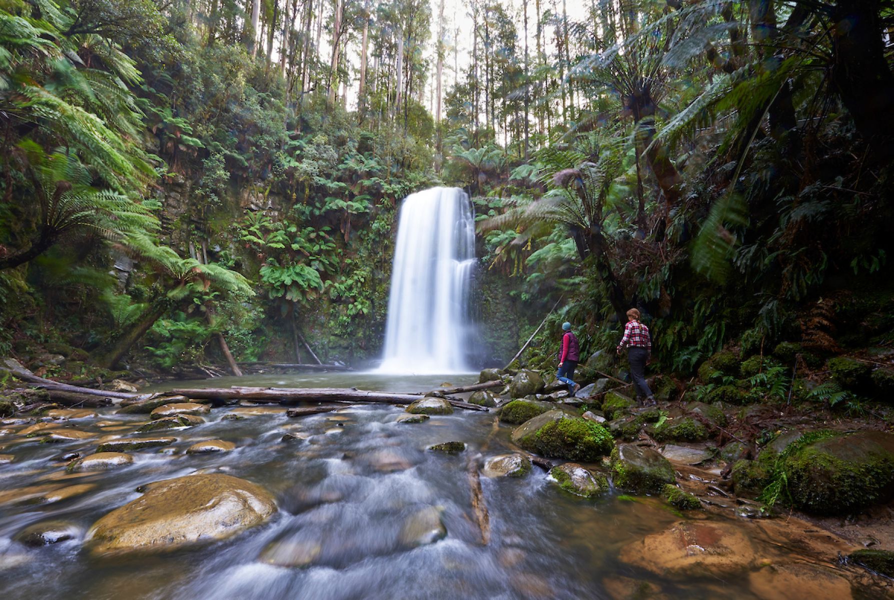 Two people walk on large stones next to a 20m waterfall