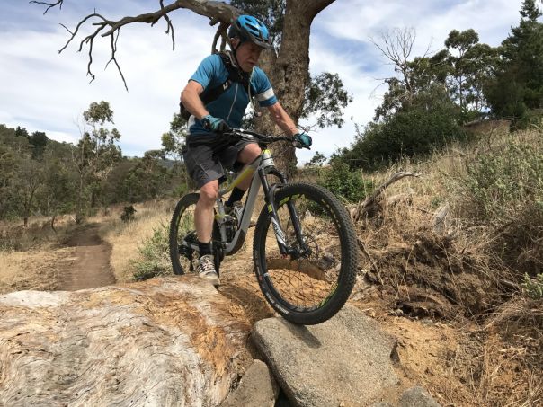 An older man riding a mountain bike across rocks and a log