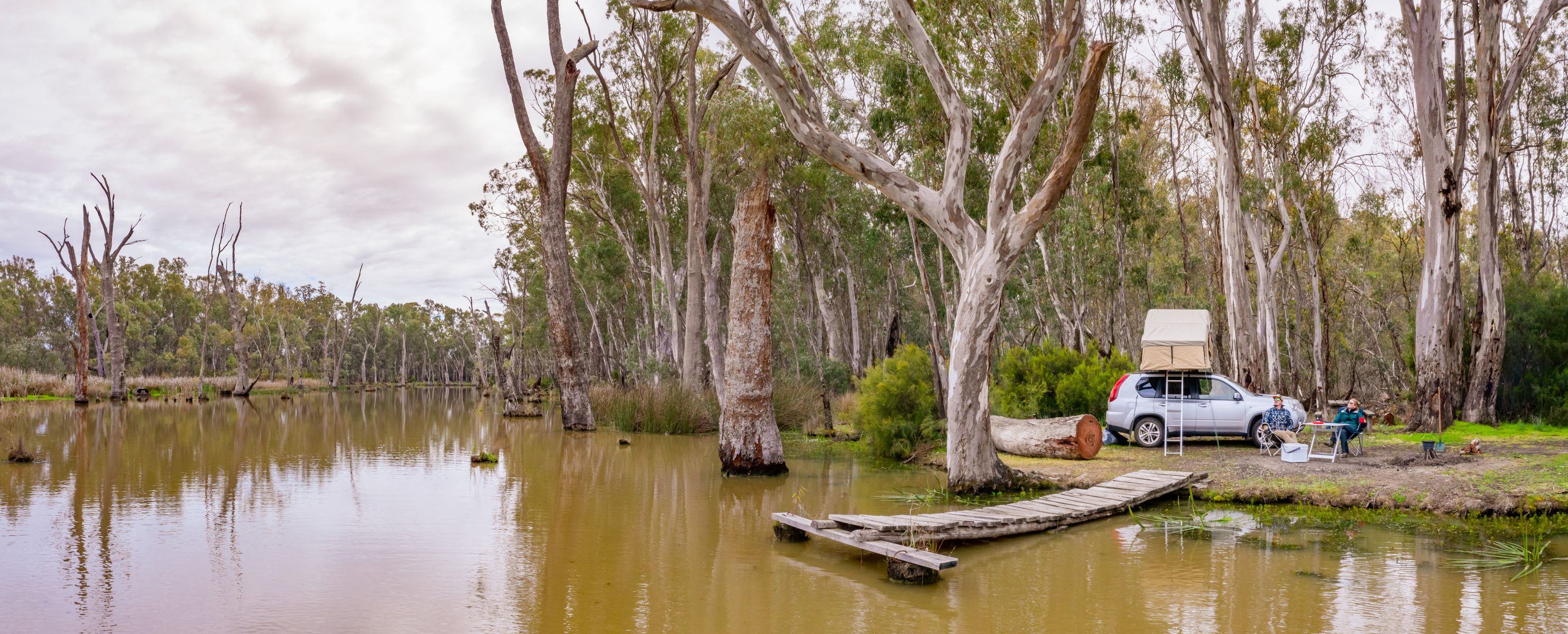 Two people camp by a river with a tent on the roof of their 4WD