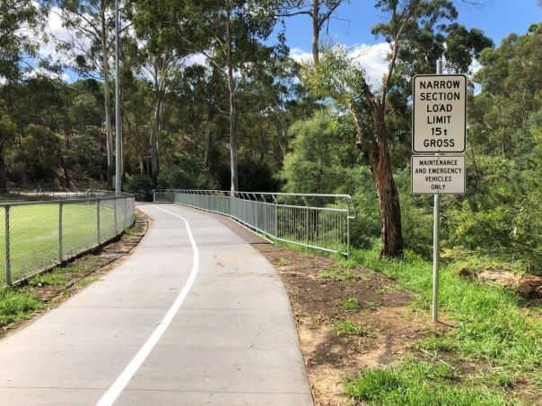 Safety road sign front right next to divided asphalt trail leading to the background over a narrow bridge. Background trees