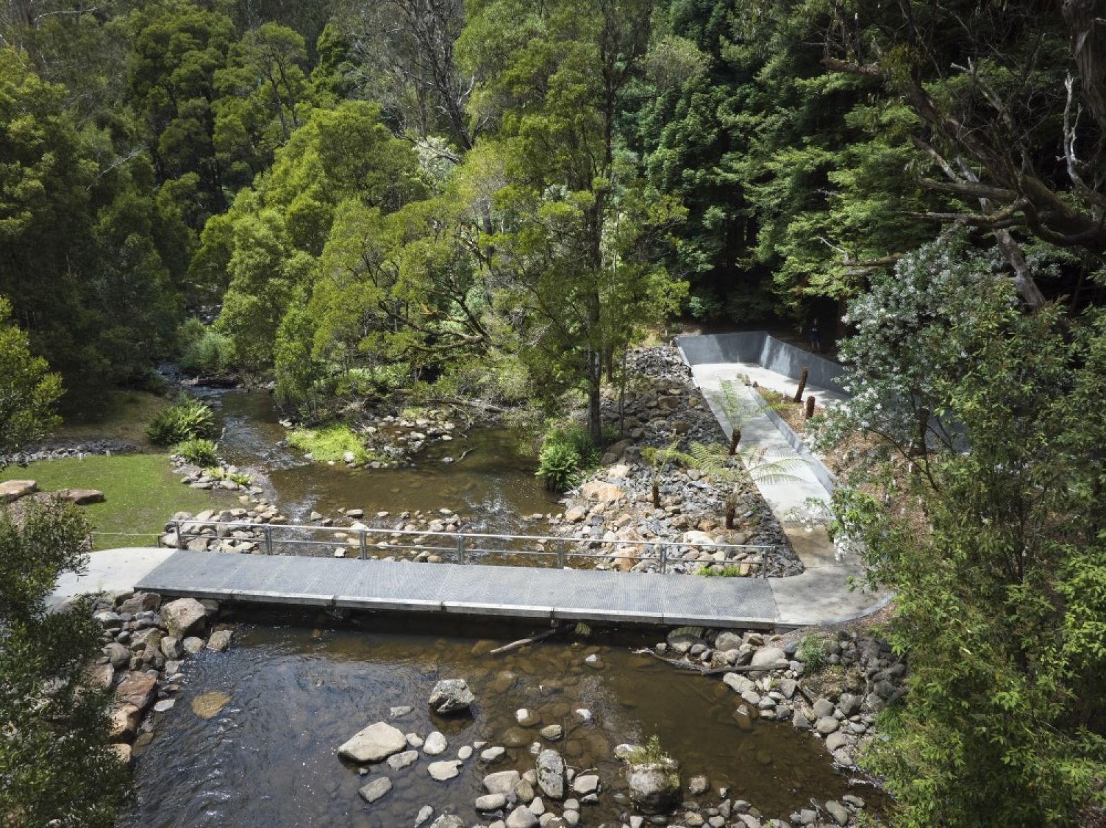A concrete and metal walkway goes over a small rocky river. A variety of green trees surround the area.
