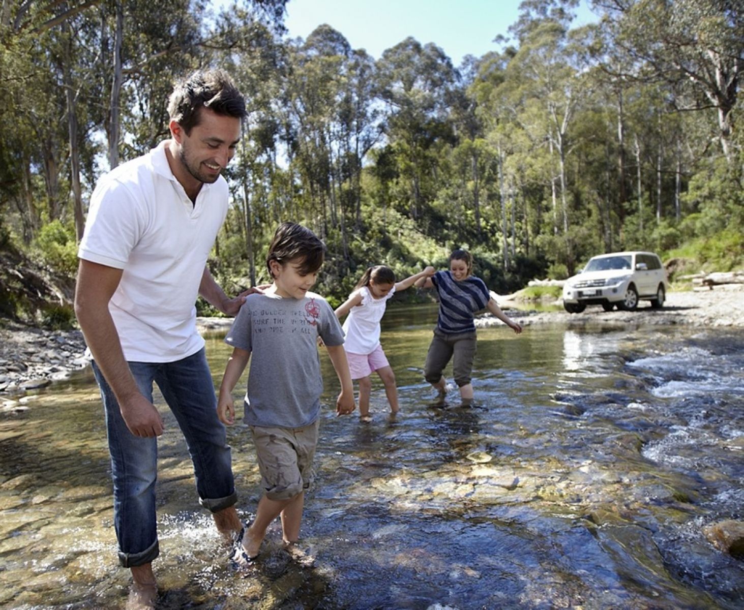 A family of four walking through a river in the forest with their 4WD in the background. 