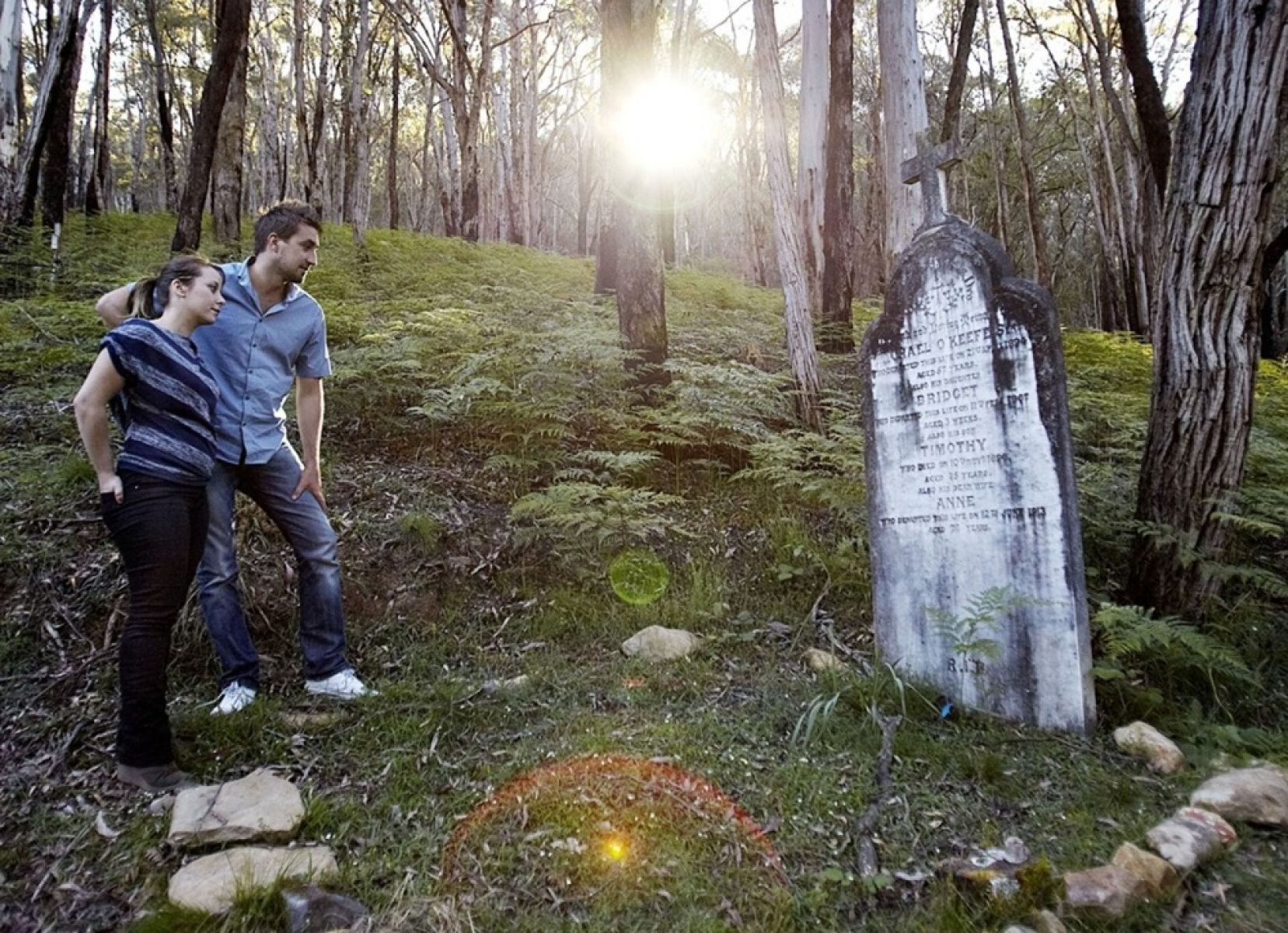 A couple looking at a tombstone in the forest surrounded by trees. 