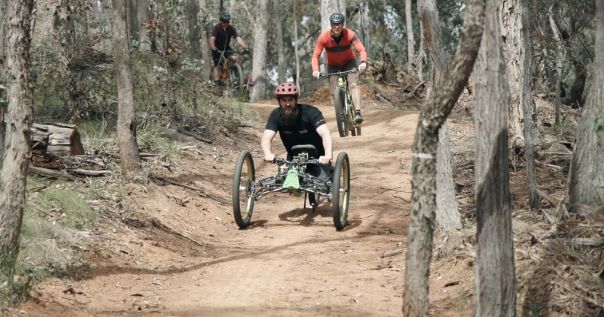 Two people riding on a gravel mountain bike trail, with one bike being a low 3 wheel bike suitable for people with disabilities