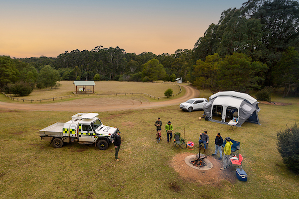 Preparing to camp in Victoria A group of friends camping in the bush. A fire pit and tent can be seen behind them.