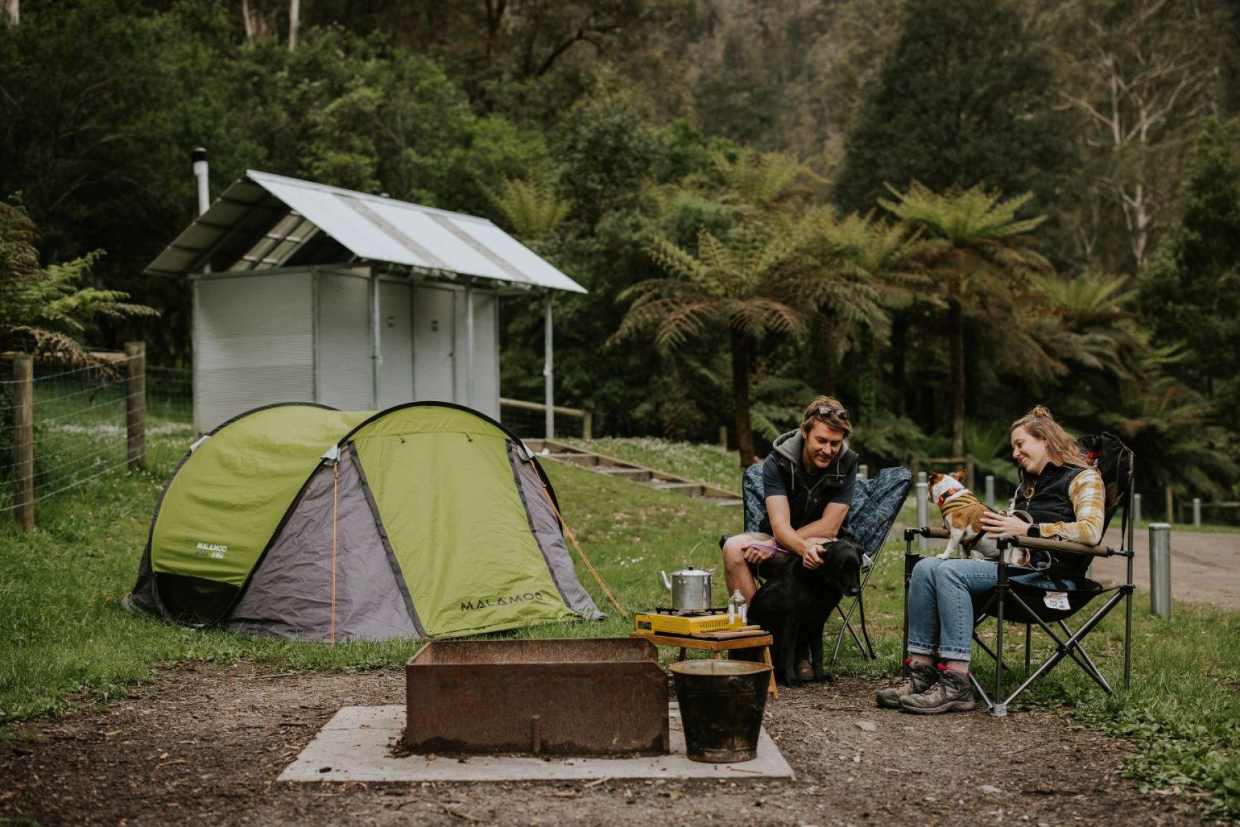 Two people sitting next to a tent in the foreground and an amenities building in background