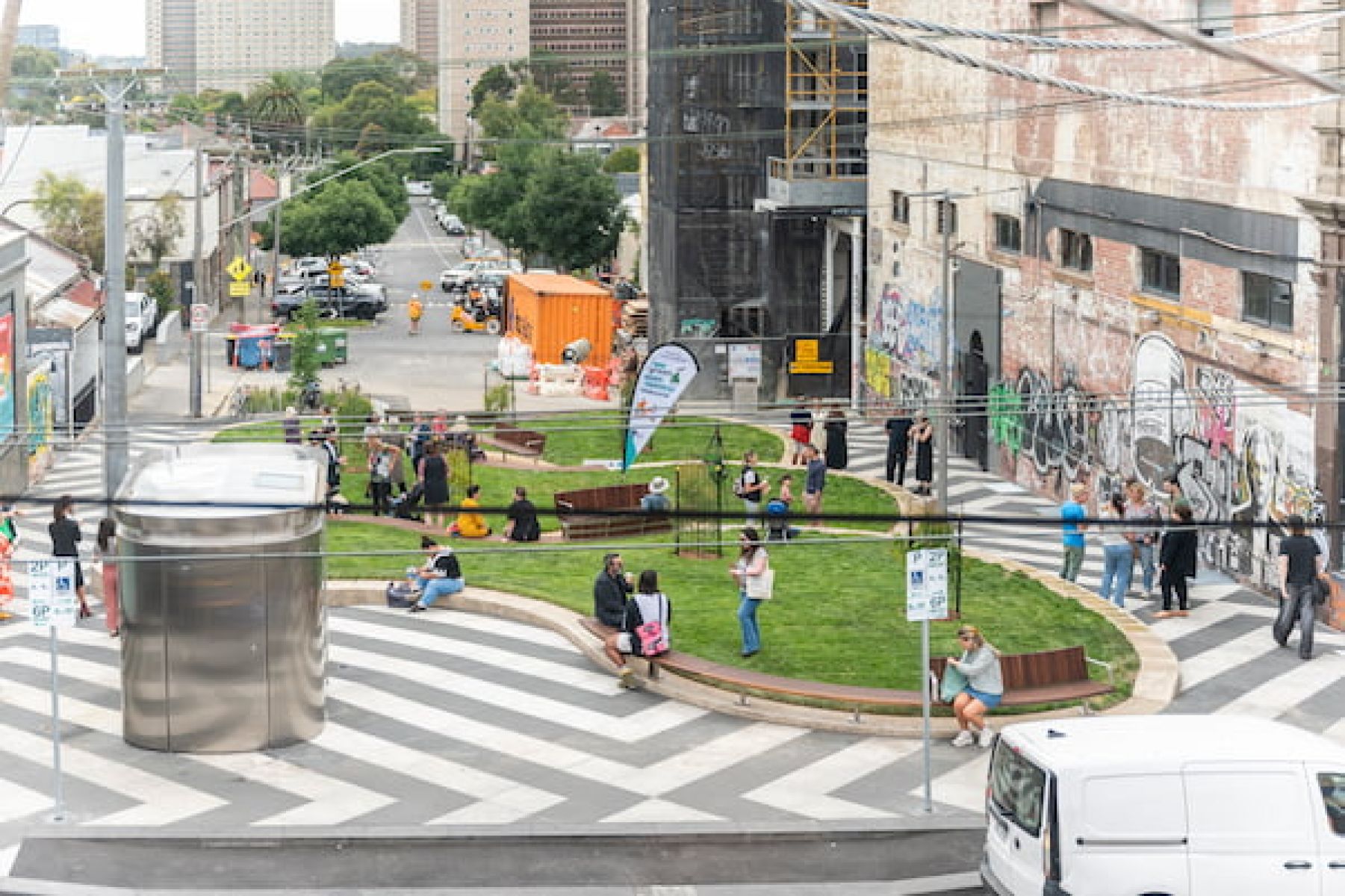 Photo of a park in a built-up urban street setting, with many people standing, sitting and walking through