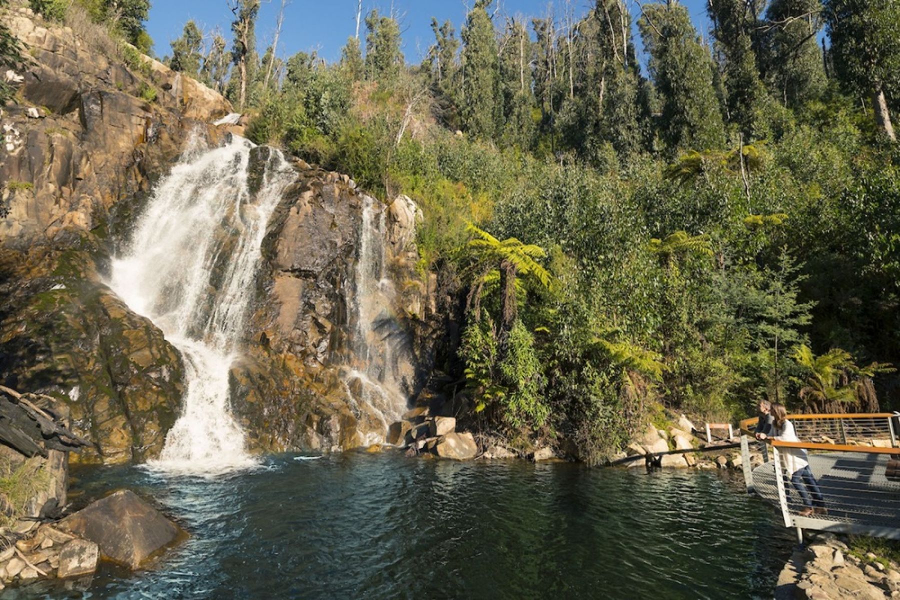 A waterfall and body of water in the forest with people standing at a viewing platform looking at the falls. 