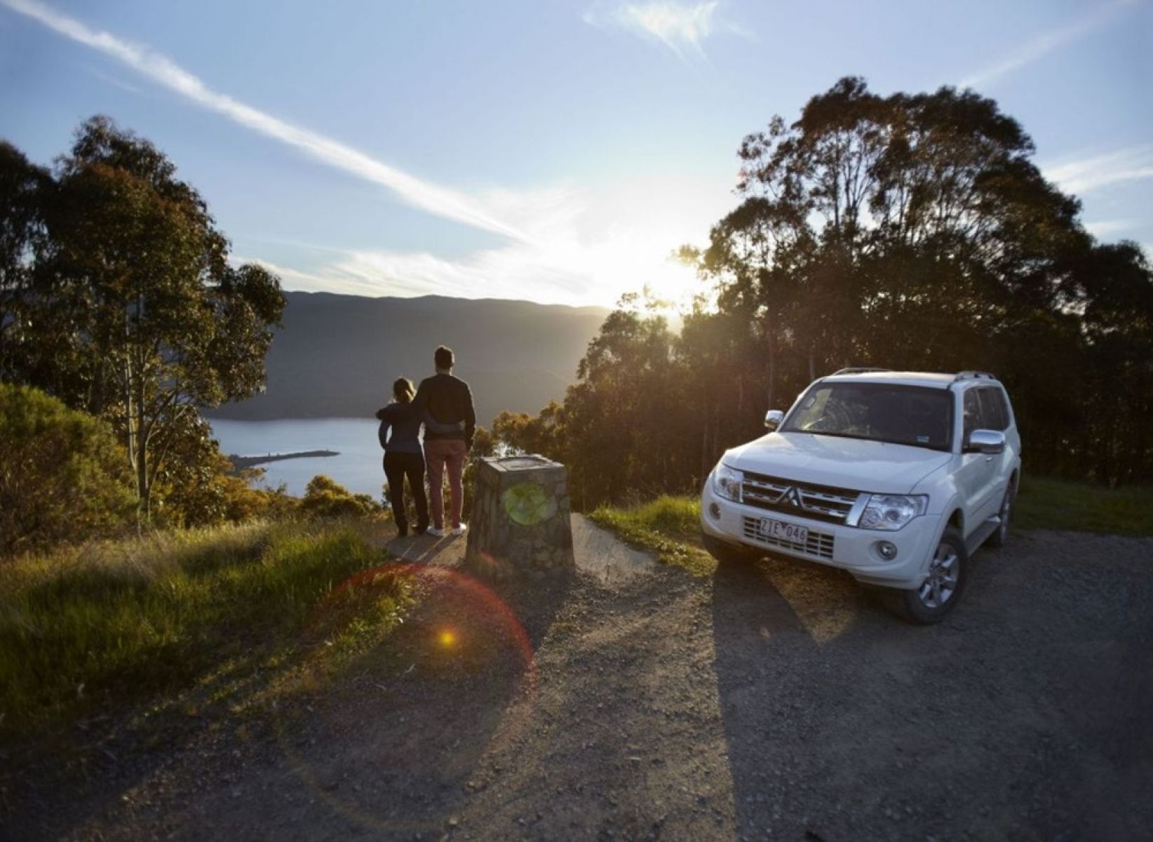 Two people overlooking a lake in the forest with their white 4WD vehicle next to them on the road. 