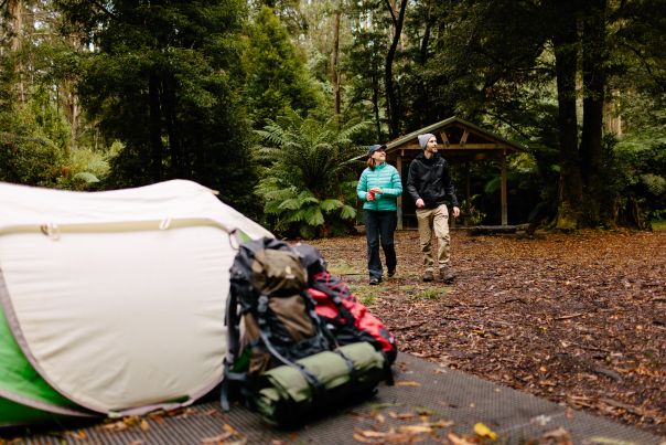 Two people walking behind a tent with overnight bags beside it