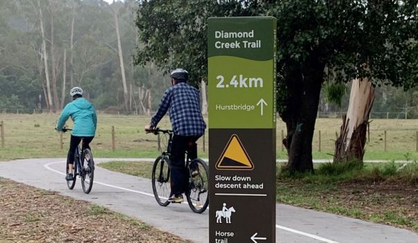 Trail sign in foreground, 2 cyclists riding into the background next to a tree on the trail