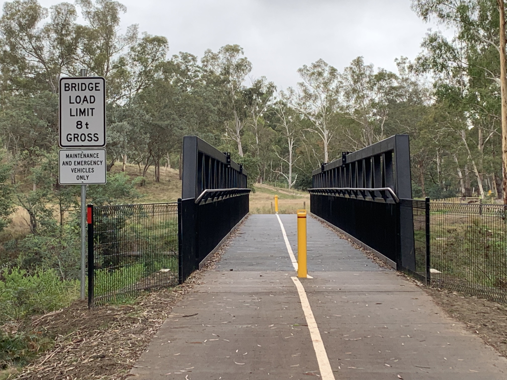 Divided trail with bridge leading to background dotted with trees. Bridge   features safety rails both sides, central yellow trail divider pole and load limit sign centre right