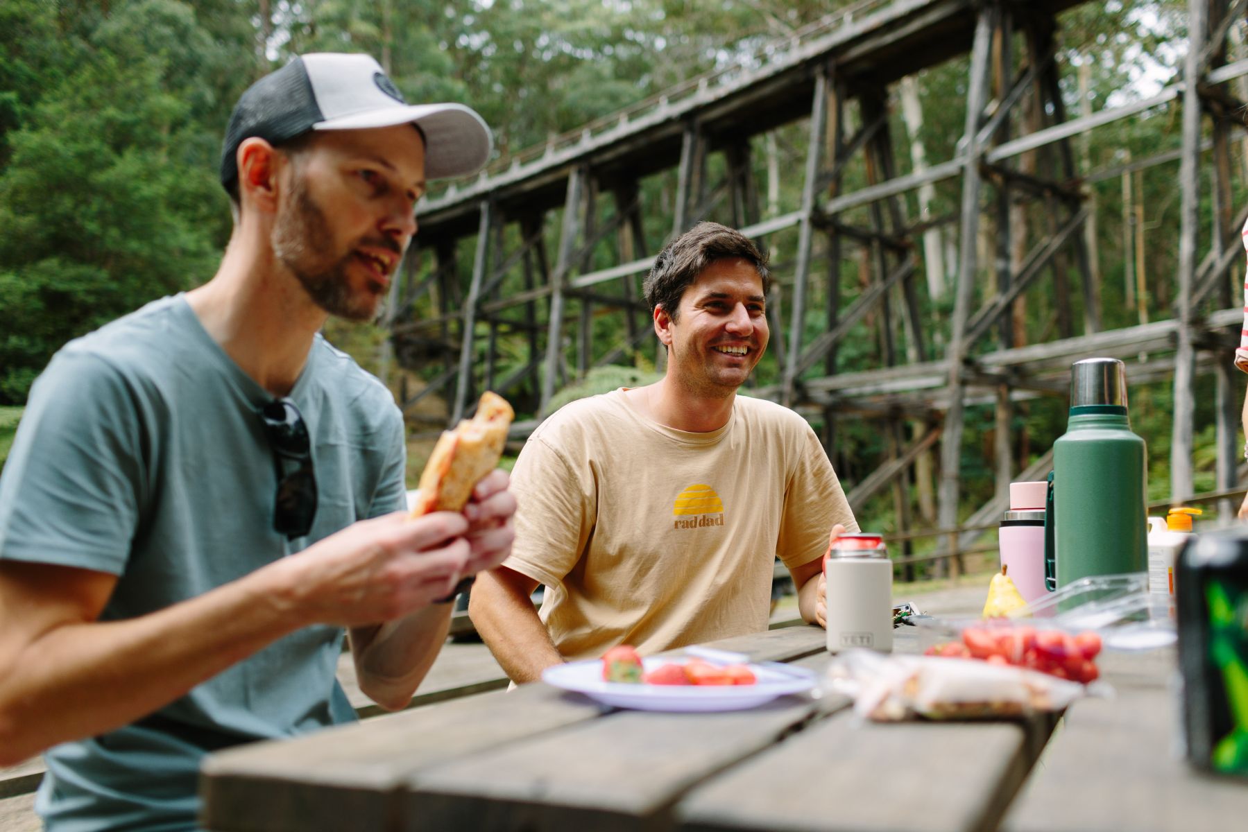 Two people eating at a picnic table with the wooden trestle bridge in the background