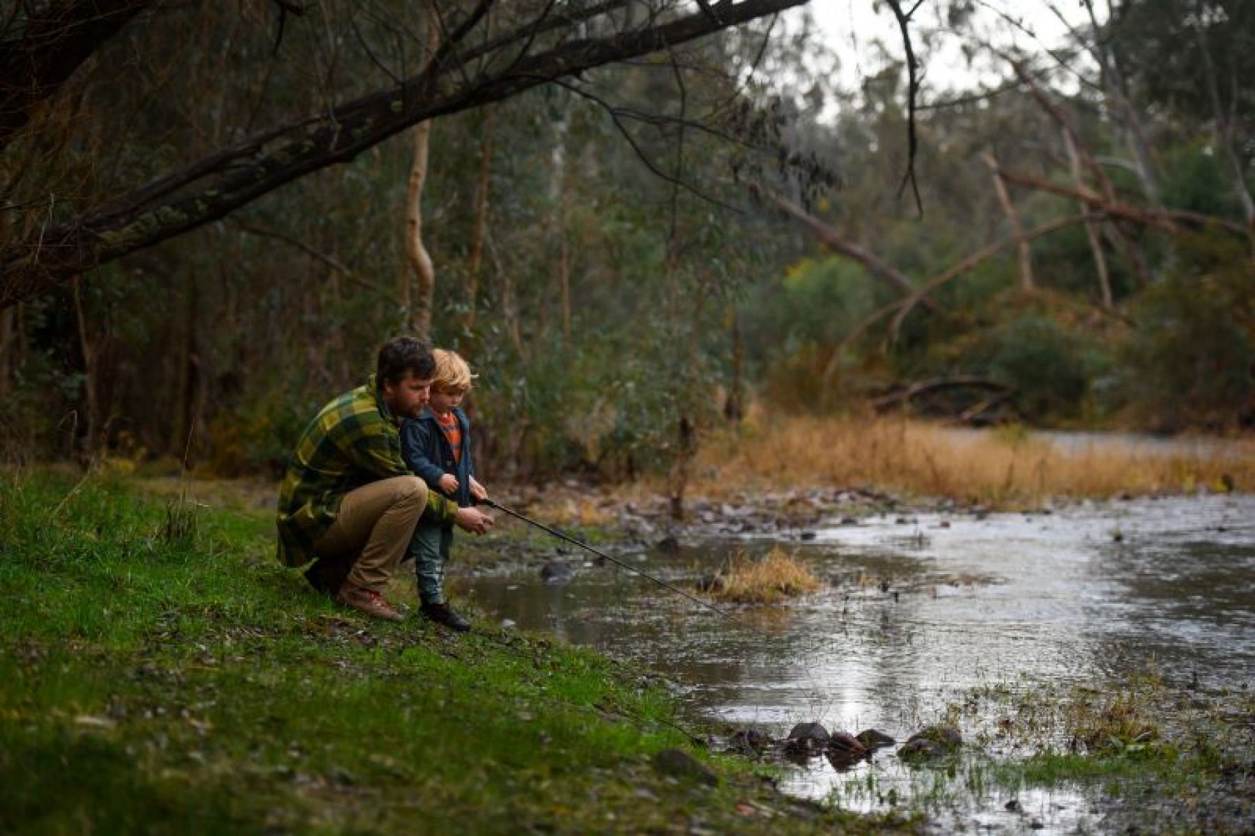 A man and child hold a fishing rod together by a river.