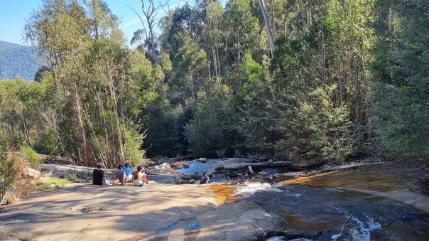 Group of people sitting down beside the top of the waterfall surrounded by forest