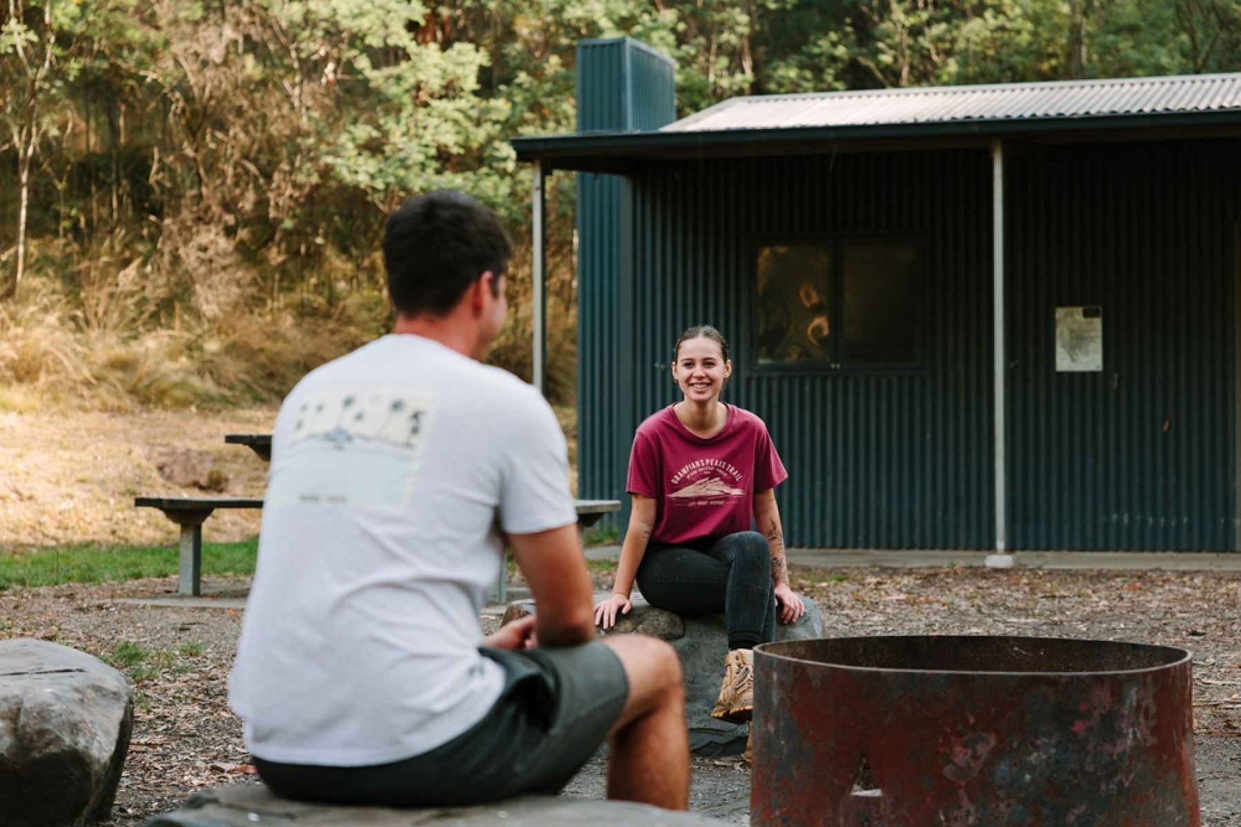 Two people sitting around a firepit in front of a hut in the forest