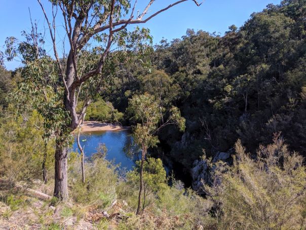 Looking down at a blue lagoon from an elevated position. Bushland surrounds the area and bright blue sky can be seen in the background.