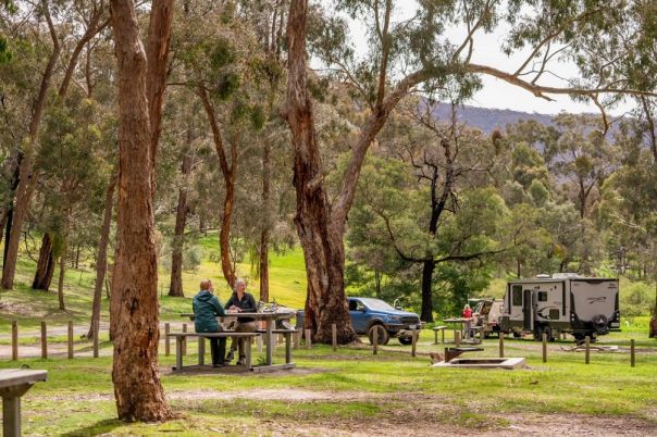 Two people sitting at a picnic table with two cars and a caravan in the background surrounded by forest.