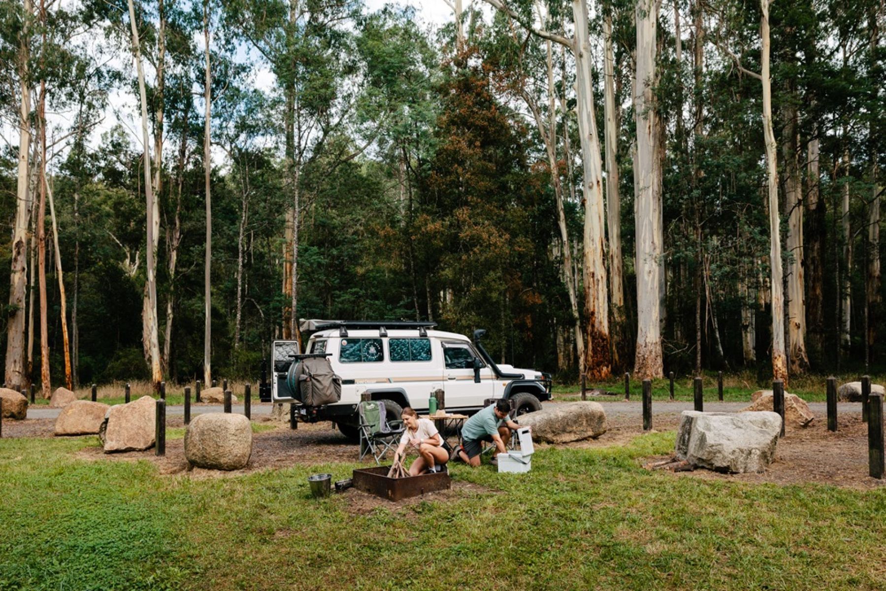 Two people at a grassy campsite with their 4wd in the background, tending to a fire and taking something out of an esky.