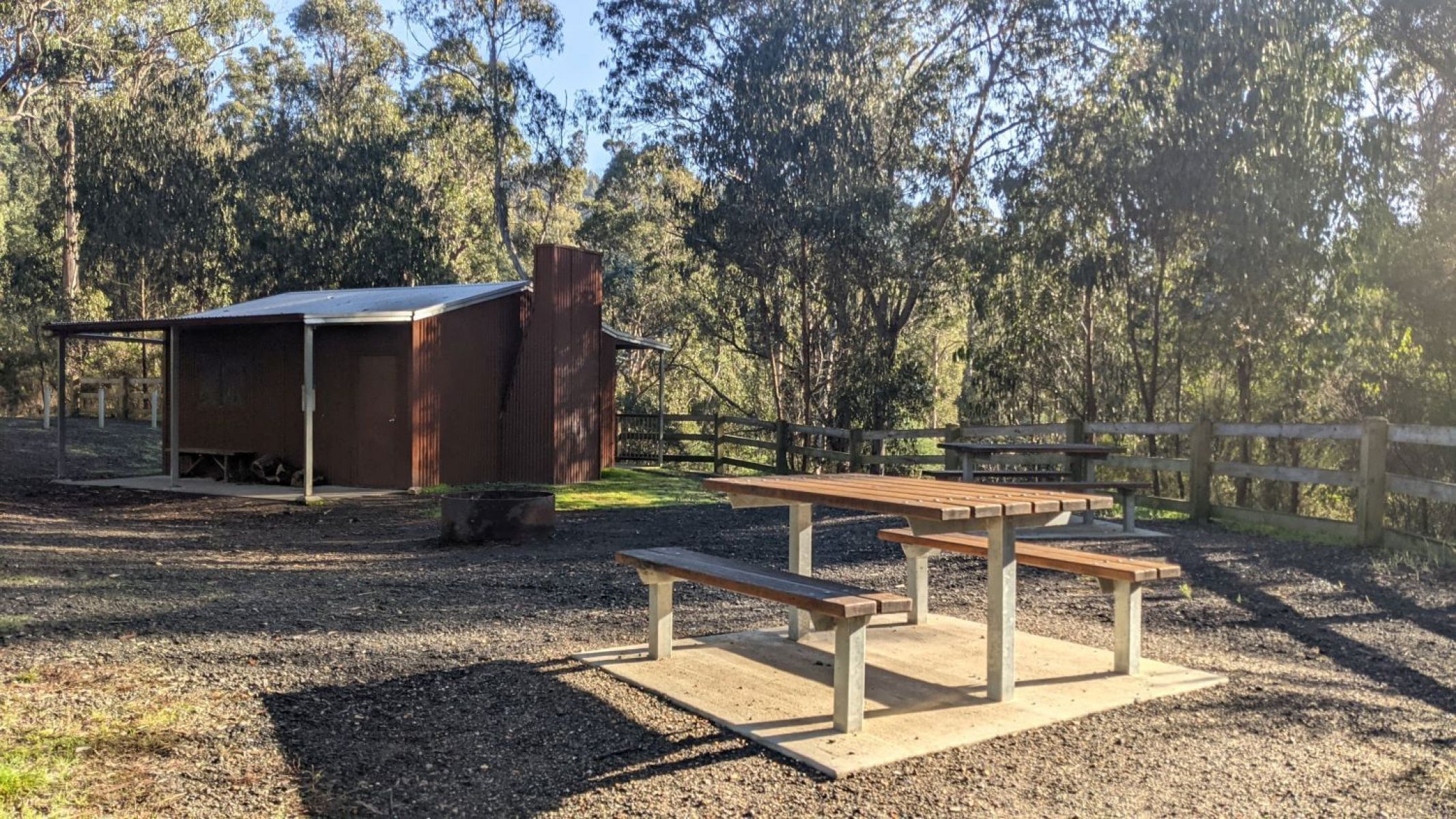 A wooden picnic table is in the foreground with a restored bush hut in the background.