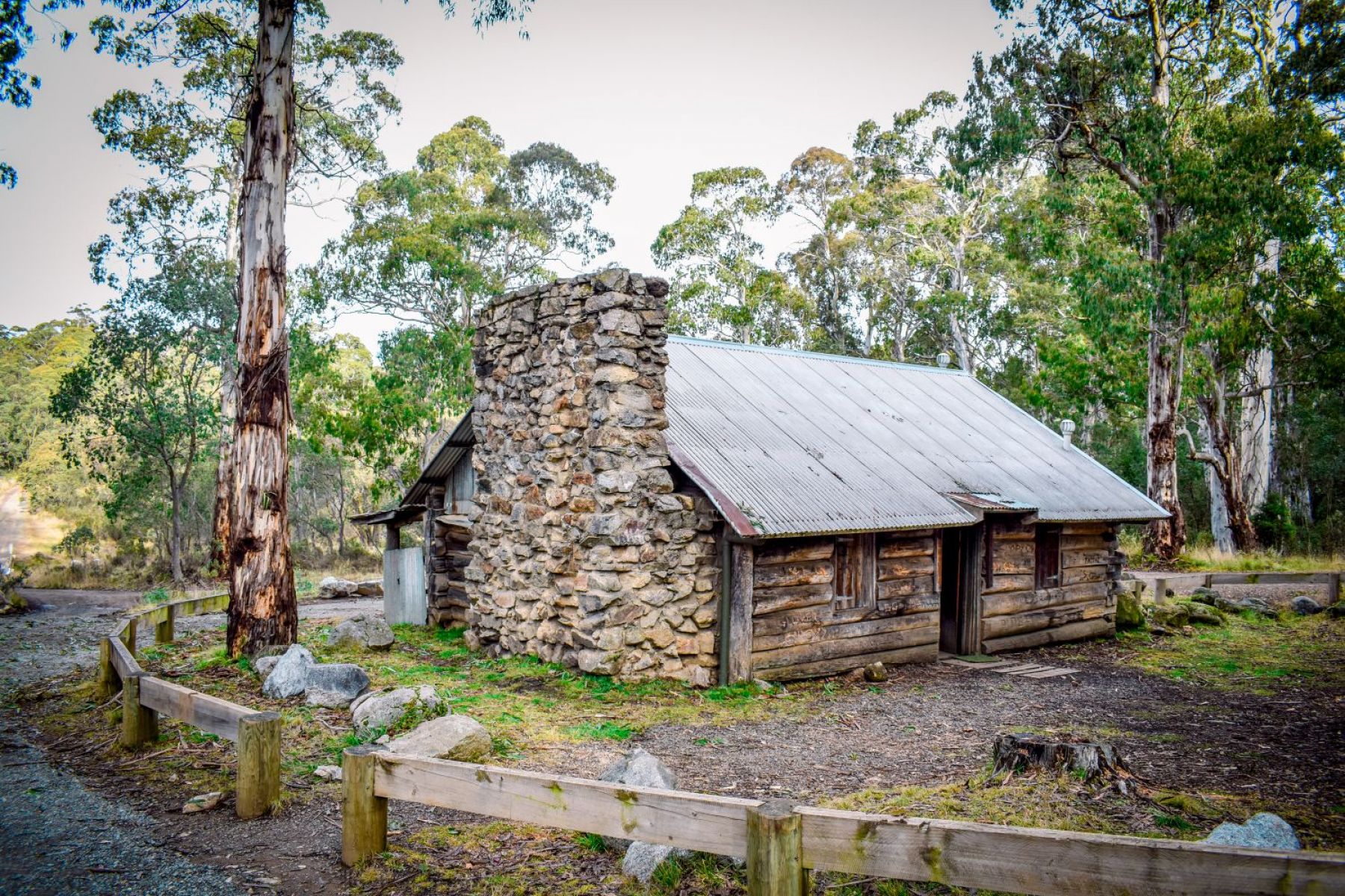 A log hut with a large stone chimney surrounded by trees. There is a gravel path leading into the hut and a small wooden fence around it. 