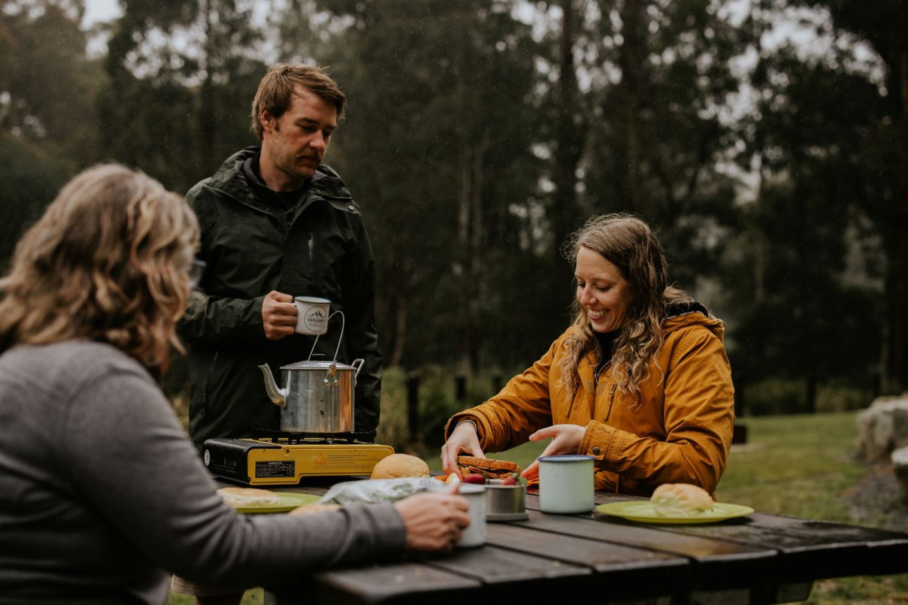 Three people enjoying lunch in the forest at a picninc table.