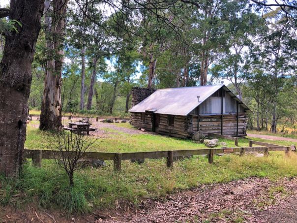 An old log hut with a metal roof is surrounded by gum trees. A wooden picnic table sits outside the hut and it is surrounded by low wooden fencing.