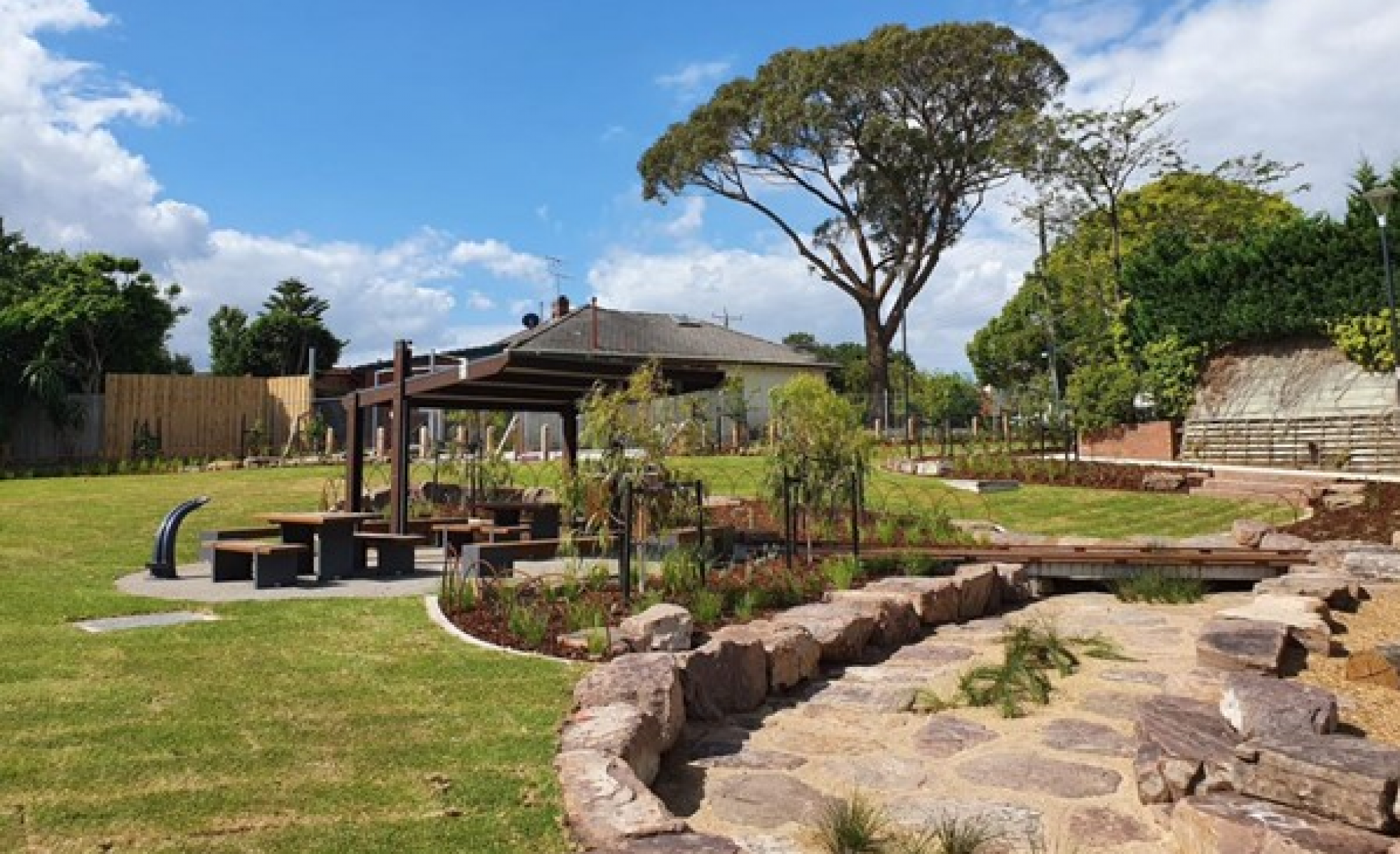 Landscaped area with rock walking path in right foreground, grassed area on left and covered open shed with table and seating in the centre