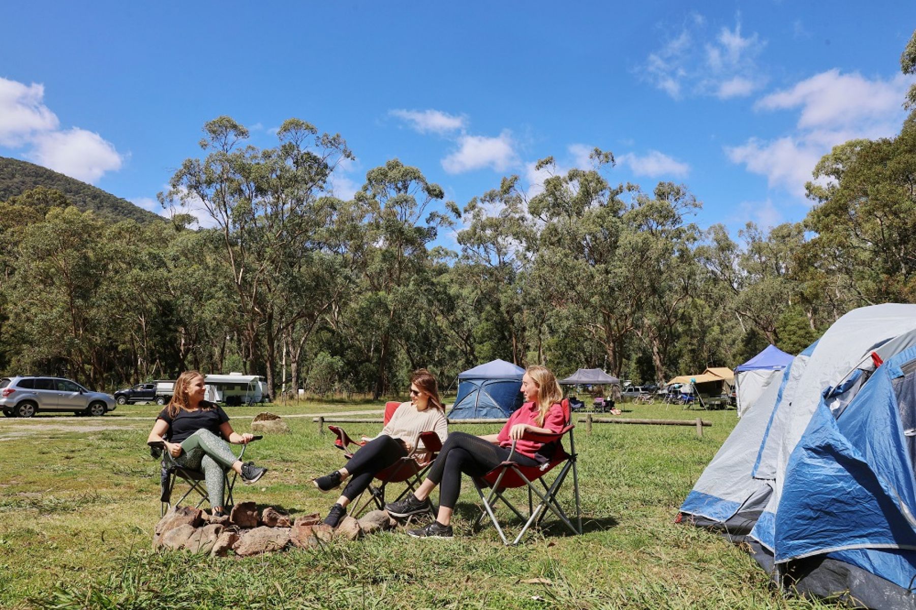 People sitting in front of tent in grassy clearing surrounded by forest