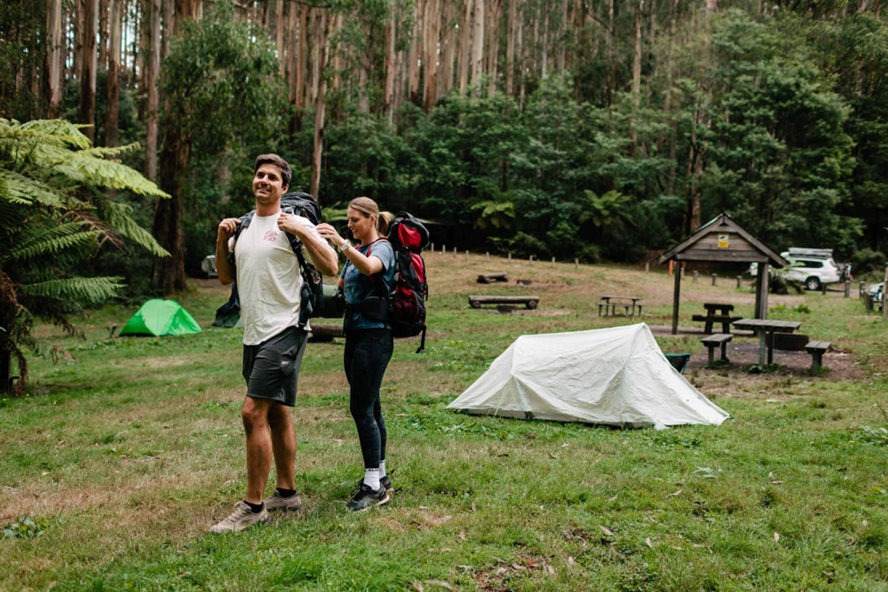 Two people at a forest campground. One person is helping the other put a packpack on. 