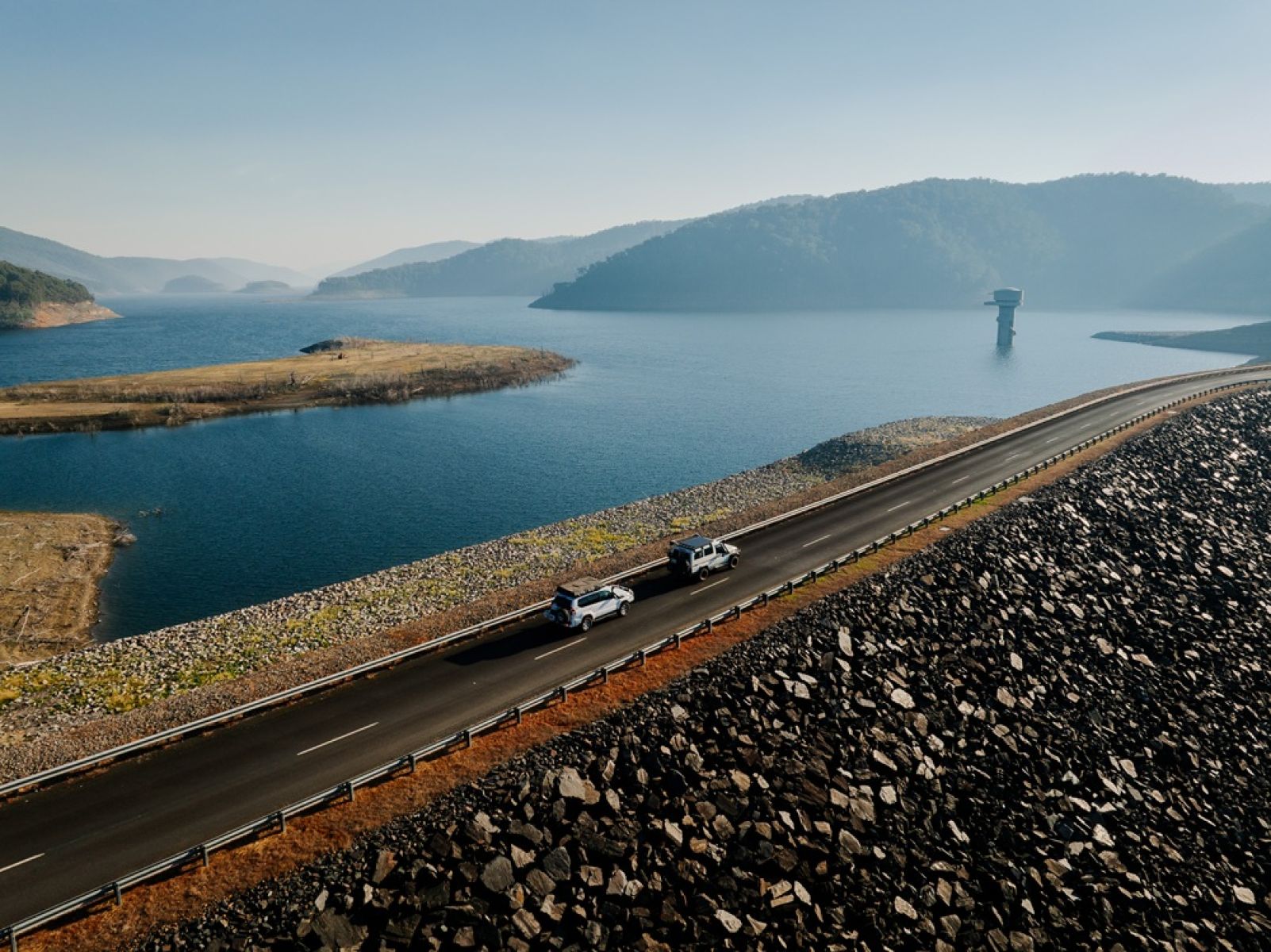 Two 4WD vehicles crossing a dam wall with a large dam surrounded by mountains in background