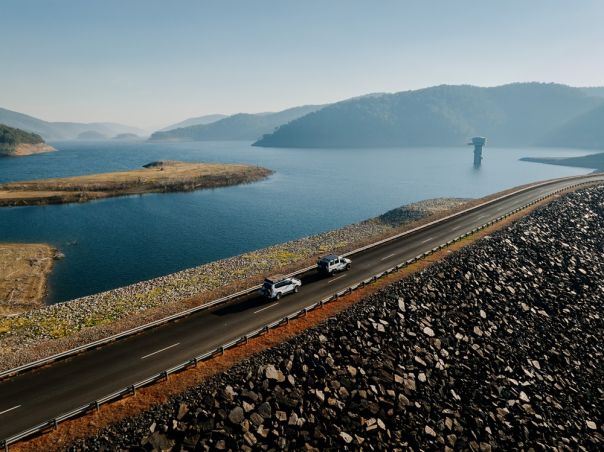 Two 4WD vehicles crossing a dam wall with a large dam surrounded by mountains in background