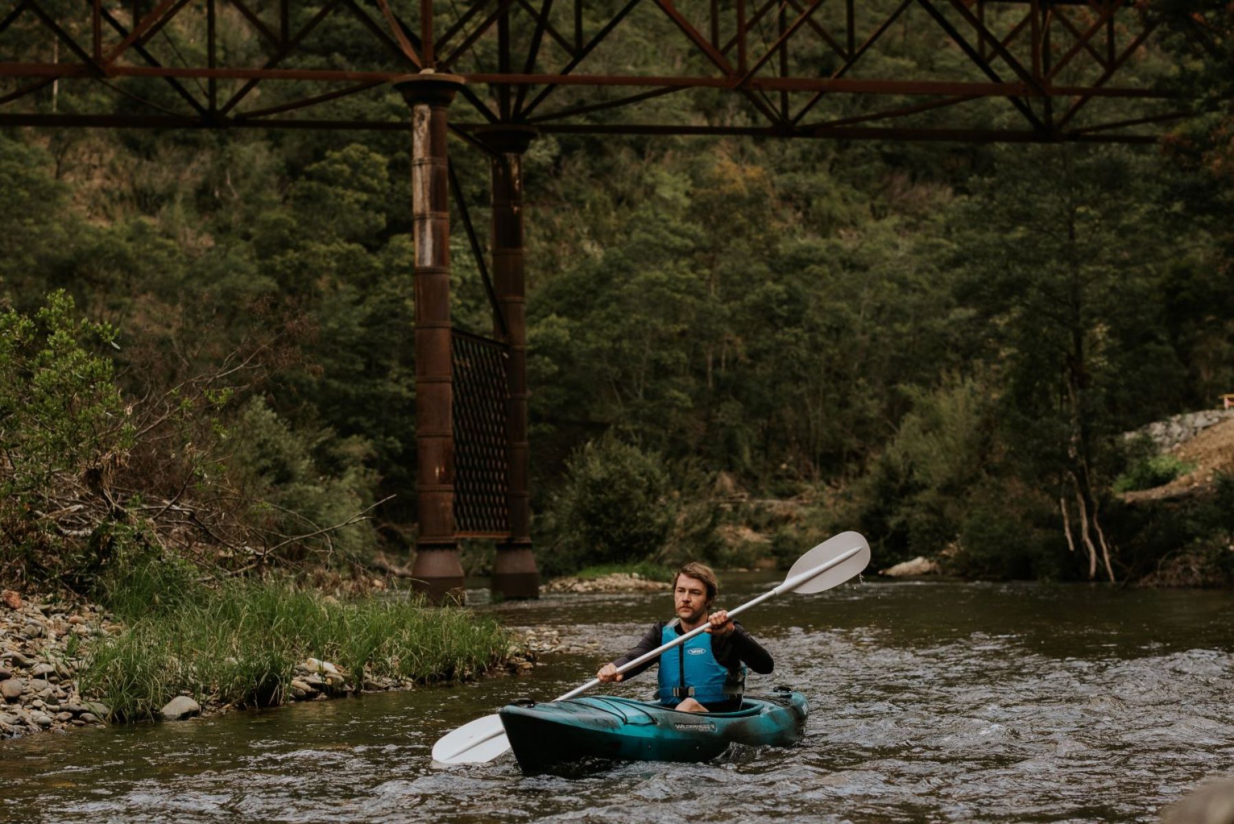 A person paddling on light rapids. A bridge is in the backrground and surrounded by trees.