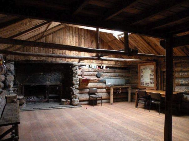 Inside an old log hut which has a stone fireplace which includes a fireplace and table and chairs.