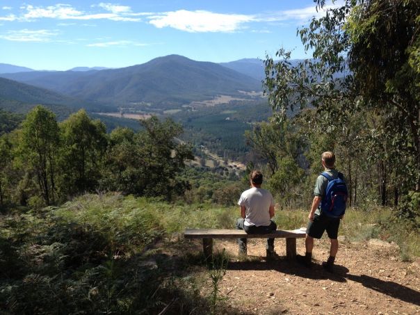One person sitting at a bench seat and another standing, overlooking the forest beyond.