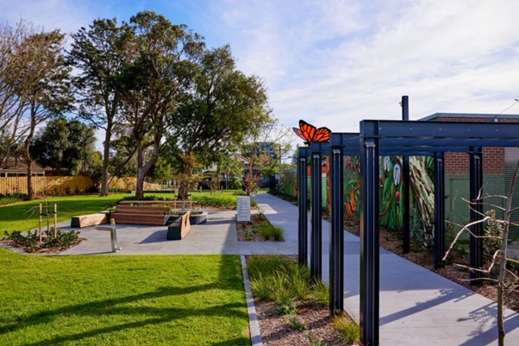 Walkway with trellis shading on right, green space and seating on left, trees in background