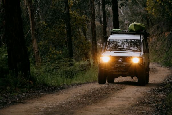 A white 4WD driving with headlights on a dirt road, canoes on the roof. 