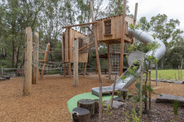 Playground with climbing frame, slide, surrounded by wooden boardwalk. Tree in foreground