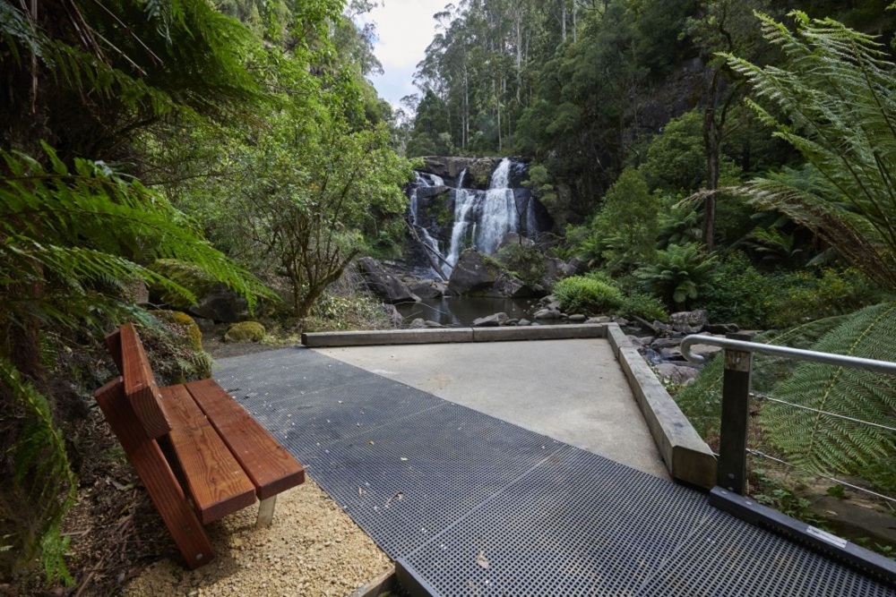 A boardwalk and wooden bench looking across to a waterfall inthe forest.  