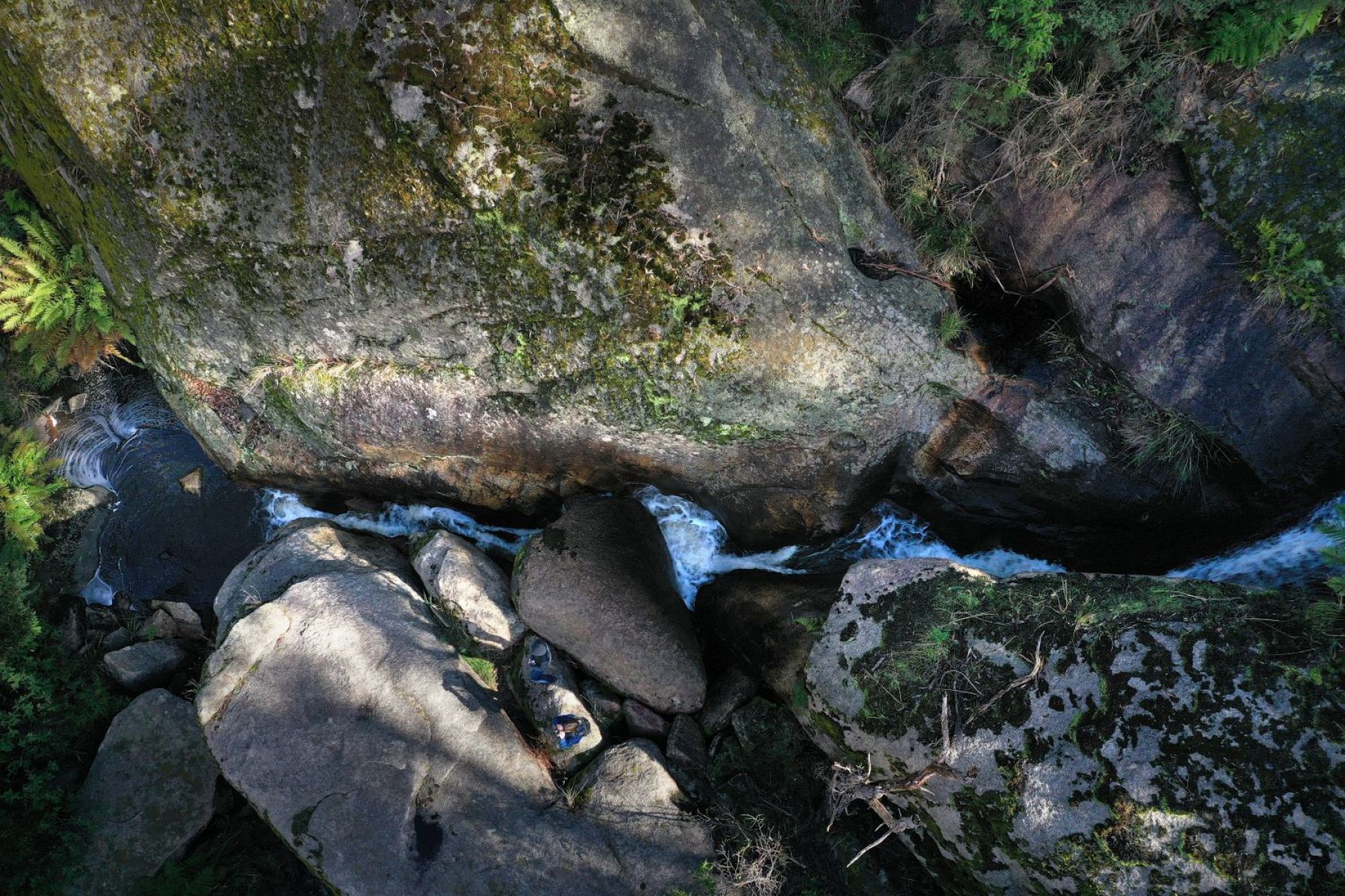 A view from above a small waterfall running between large moss-covered boulders.