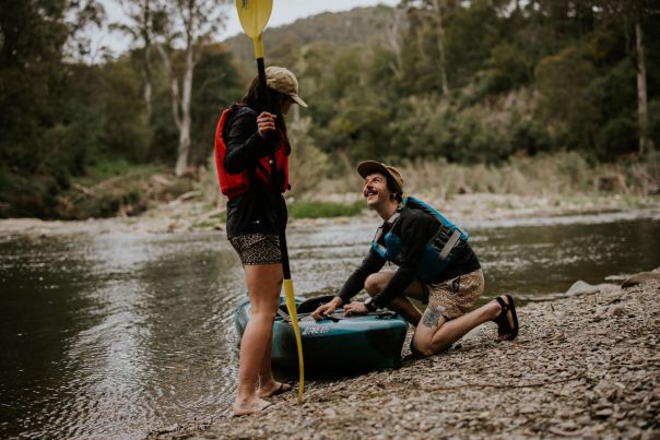 Canoe by the river, with a man kneeling and looking at a person with their back to the camera