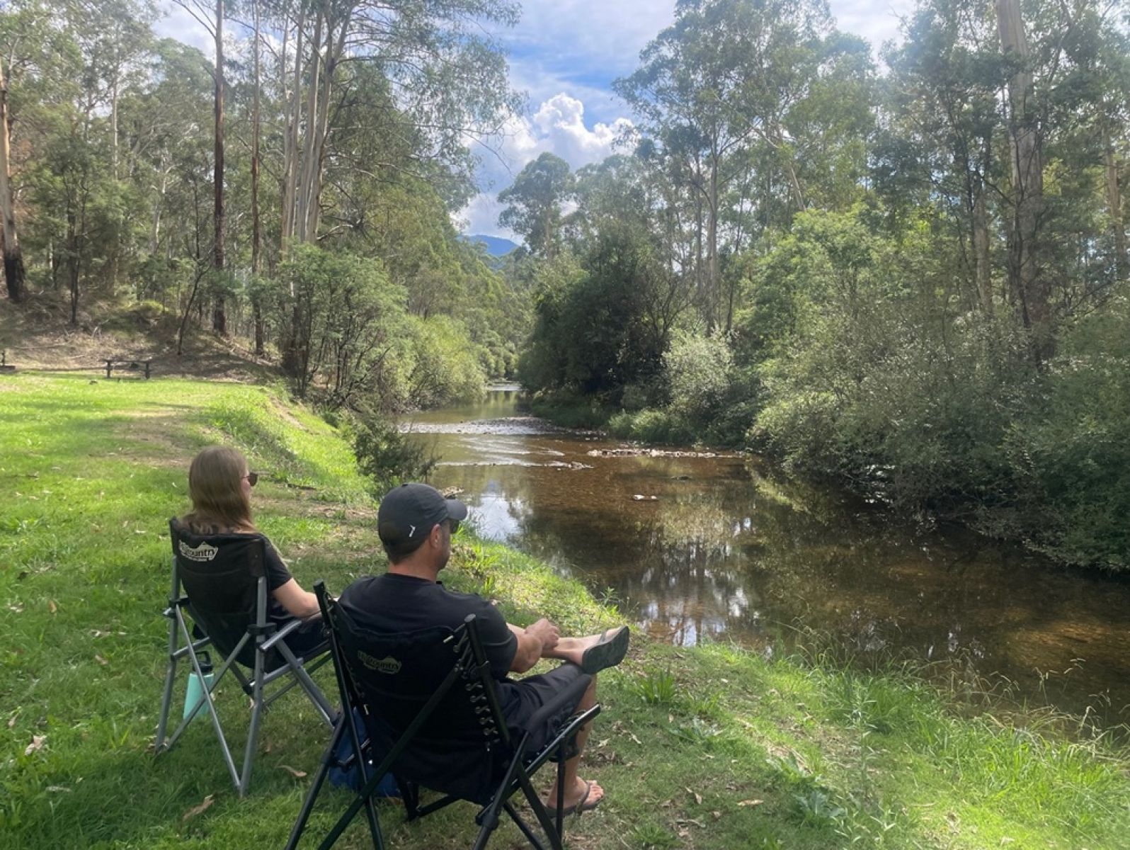 Two people sitting in chairs beside a wide shallow river surrounded by trees