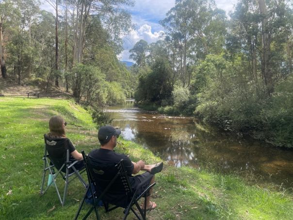 Two people sitting in chairs beside a wide shallow river surrounded by trees