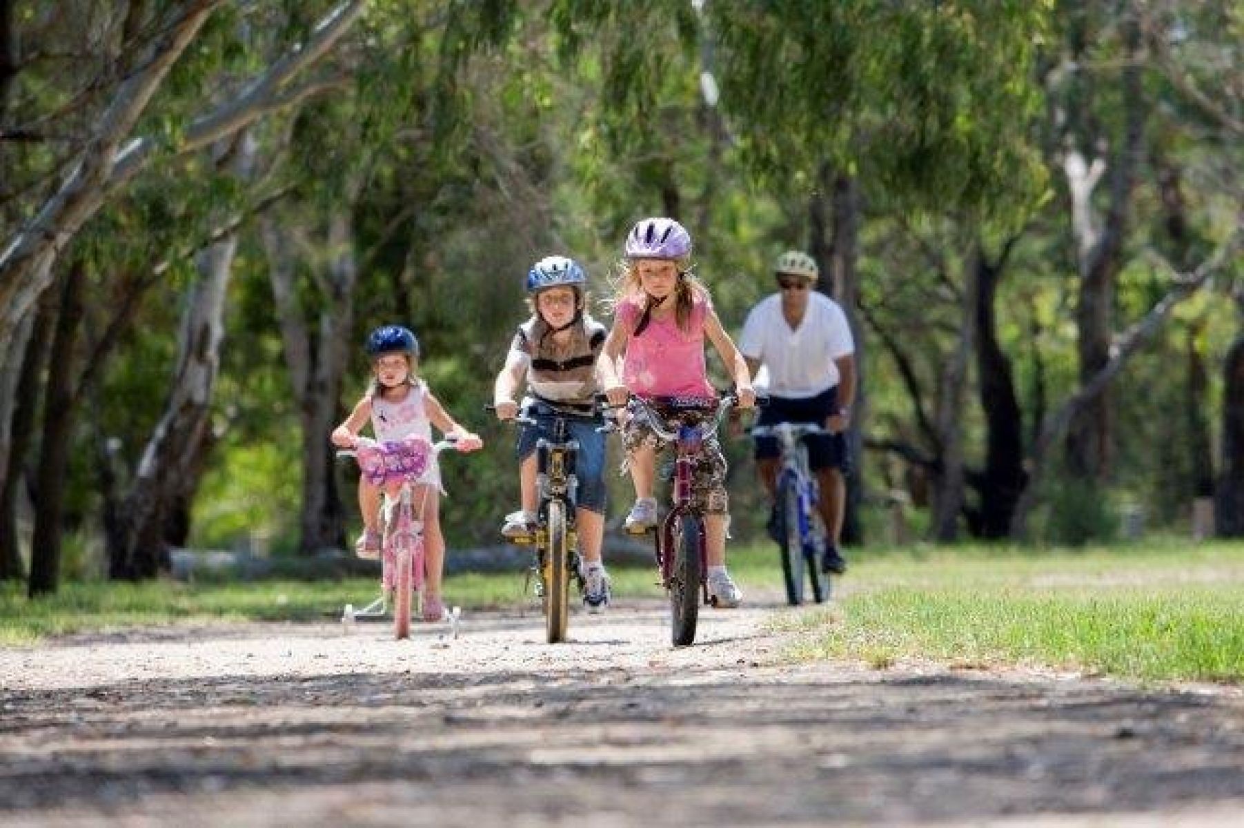 Three young people riding a bike, with a man in the background, also riding his bike on the bike path.