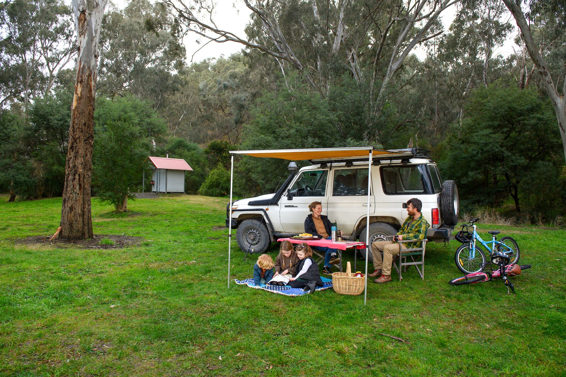 Two adults and three children sitting together smiling in front of a white vehicle at a campground. The campground has green grass and is surrounded by trees, a toilet block is in the background.